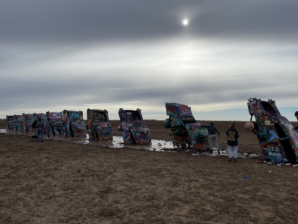 a group of people standing next to a group of tents with Cadillac Ranch in the background